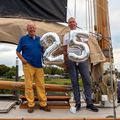 Walter Esch en wethouder Van Leeuwen op zijn schip. Ze houden beiden een zilveren ballon vast in de vorm van een cijfer: een 2 en en een 5. Samen vormen ze het getal 24. 