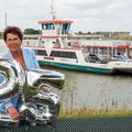 Andrea Haubrich staat op de voorgrond en houdt de zilveren ballonen vast in de vorm van het getal 25. Ze staat buiten voor de rivier. Achter haar zie je de pont in het water liggen. 