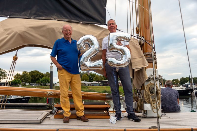 Walter Esch en wethouder Van Leeuwen op zijn schip. Ze houden beiden een zilveren ballon vast in de vorm van een cijfer: een 2 en en een 5. Samen vormen ze het getal 24. 