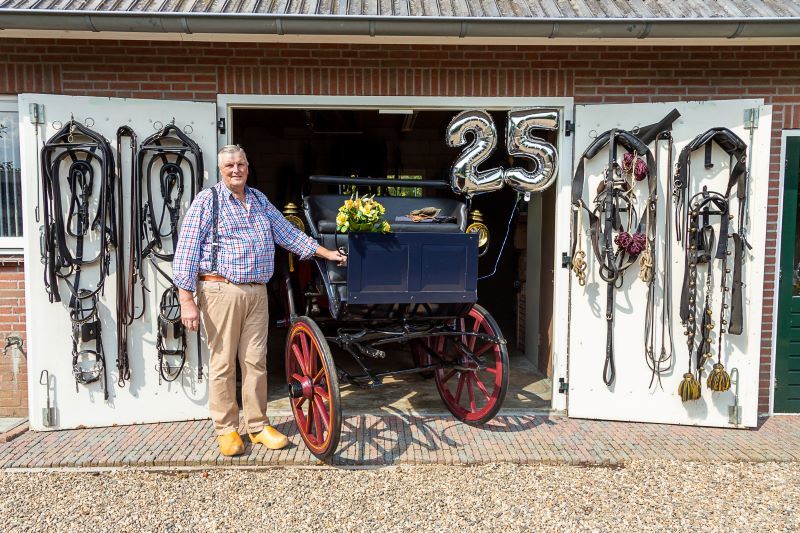 Koos Tromp staat naast zijn menwagen voor zijn schuur. Aan de deuren hangen meerdere tuigen. Rechts boven zweven de zilveren ballonnen in de vorm van het getal 25