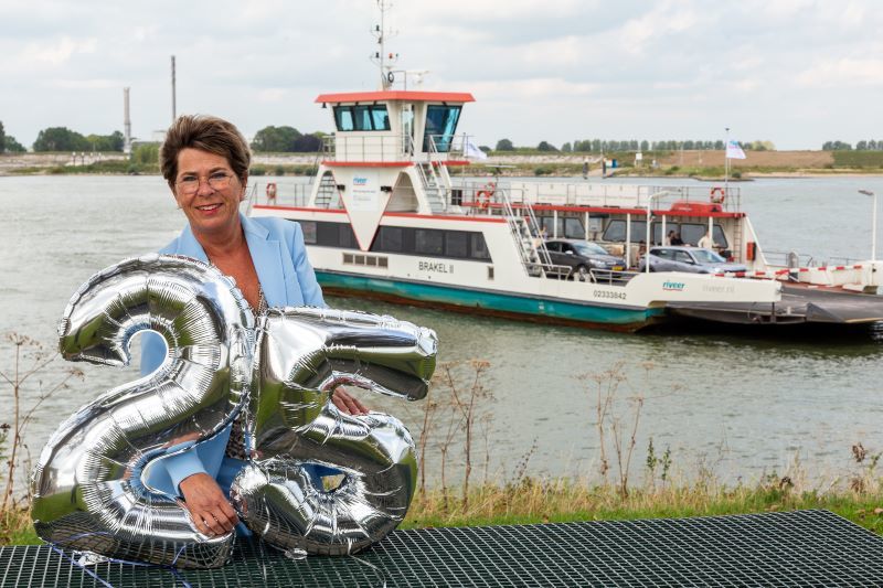 Andrea Haubrich staat op de voorgrond en houdt de zilveren ballonen vast in de vorm van het getal 25. Ze staat buiten voor de rivier. Achter haar zie je de pont in het water liggen. 