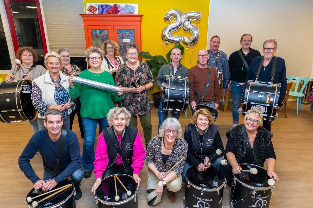 Leden van de band de Bommeleros met muziekinstrumenten. Op de achtergrond zilveren ballonnen in de vorm van het getal 25.